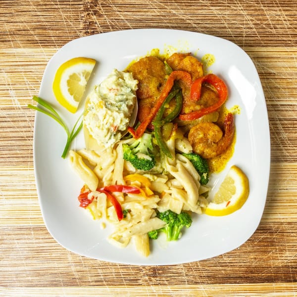 A white plate with shrimp, pasta, broccoli, bell peppers, and potato salad, garnished with lemon.