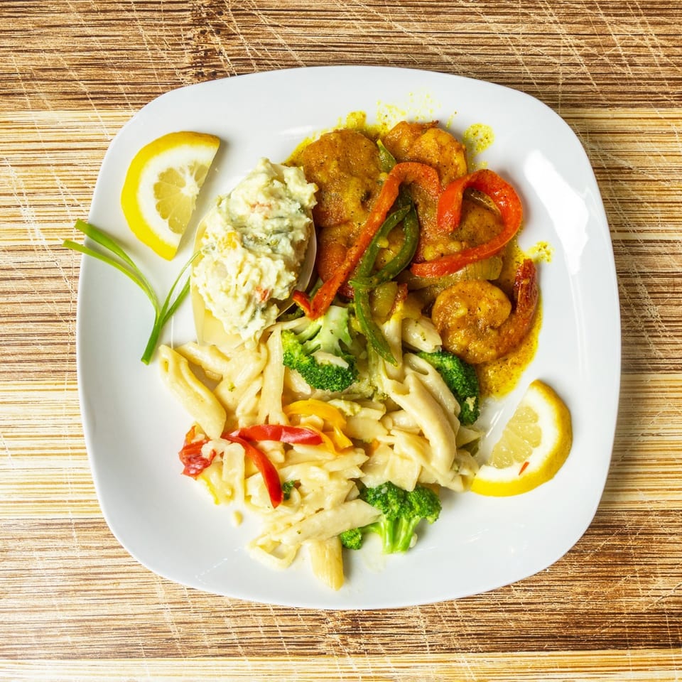 A white plate with shrimp, pasta, broccoli, bell peppers, and potato salad, garnished with lemon.