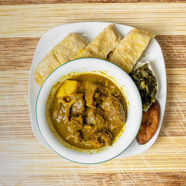 Curried meat and potatoes in a bowl, with roti, collard greens, and fried plantain on a plate.
